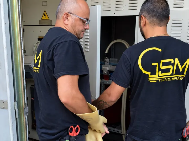 Due tecnici in maglia G.S.M., coi guanti isolanti, davanti alla cella di media tensione di una cabina